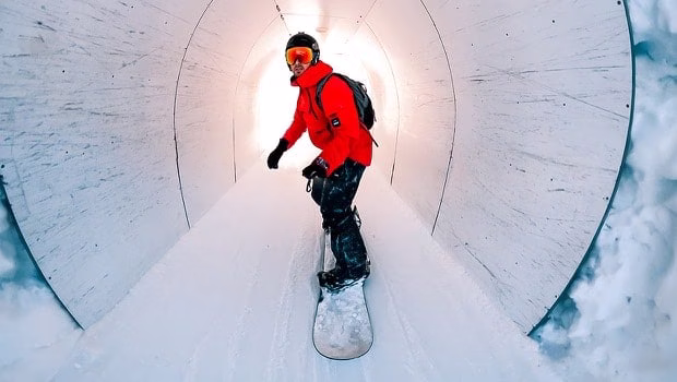 一位身穿亮红色滑雪服的滑雪者滑过覆满雪的隧道，并回望镜头。隧道后方被灯光照亮，营造出一片耀眼的白色背景。 