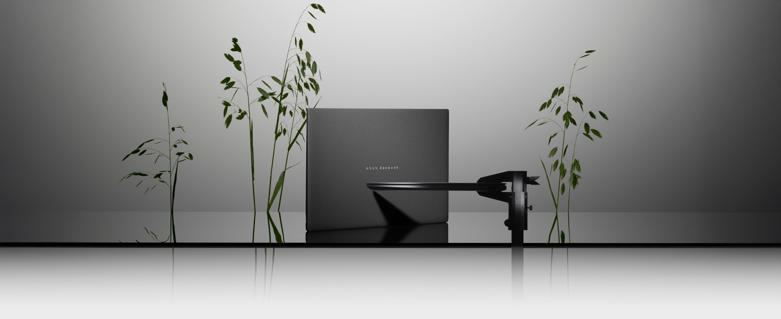 The image showcases a closed ASUS Zenbook S16 laptop on a reflective surface. A caliper beside it emphasizes the laptop’s slim profile. Green plant stems frame the scene, adding a natural contrast to the sleek, metallic design. 