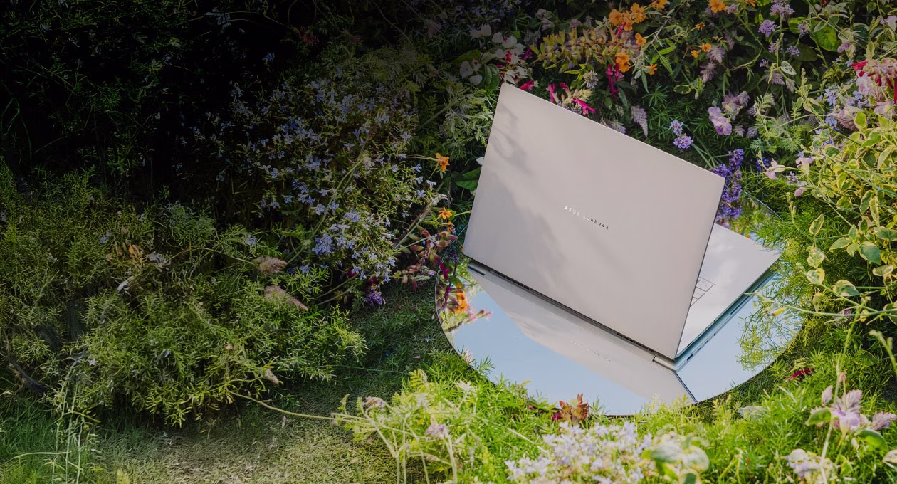 A sleek laptop rests on a circular mirror amid a vibrant garden, reflecting its immersive 3K OLED visuals and cinematic sound.