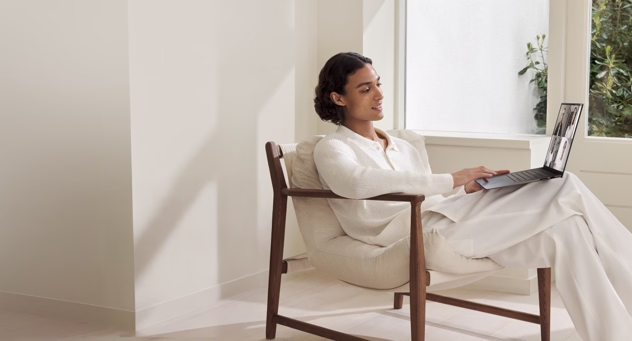 A person in white attire sits in a bright, minimalist room, engaged in a video call on their laptop. 