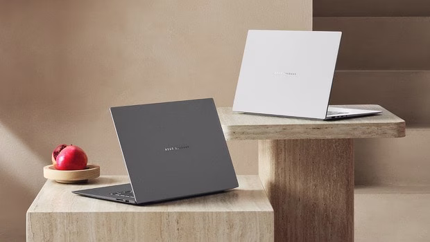 Two ASUS Zenbook S16 laptops in contrasting colors are elegantly displayed on stone pedestals in a minimalist setting. 