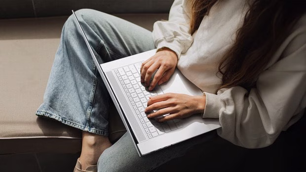 A person in casual attire types on a laptop while seated comfortably on a cushioned surface.
