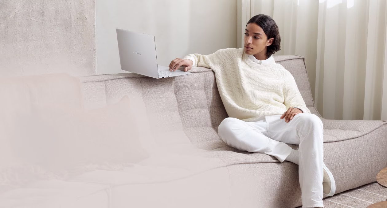 A person in white sits on a beige sofa, casually using a silver laptop placed on the armrest. 
