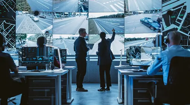 控制室里有两人正在分析多个监控屏幕显示的交通画面。环境忙碌而专注，充满高科技氛围。