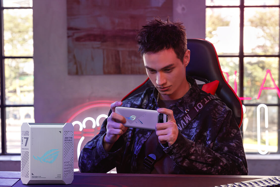 一名男子坐在游戏椅上,手持ROG手机进行游戏,ROG 魔盒 Air WiFi 7路由器置于他面前的桌面上,整个游戏室沐浴在日光中。