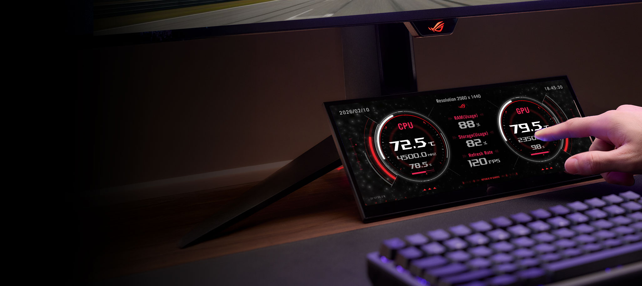特写镜头显示,一根手指正触摸着ROG XG129C的副显示屏,屏幕上显示着红色的圆形电脑性能指示器,该显示屏位于桌面上的机械键盘后面。