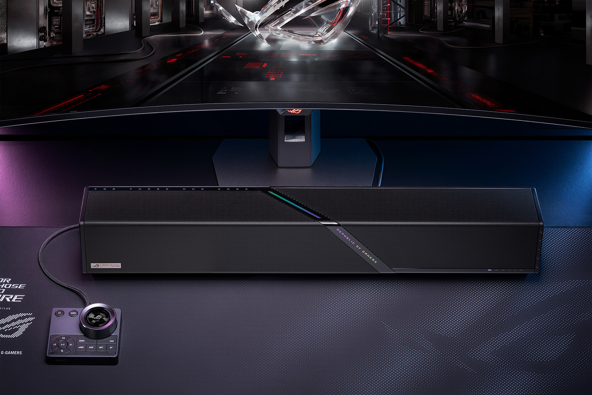 A close-up shot of a ROG 加拉尔soundbar positioned directly beneath a monitor. To the left, an all-in-one audio control hub sits on a mousepad.