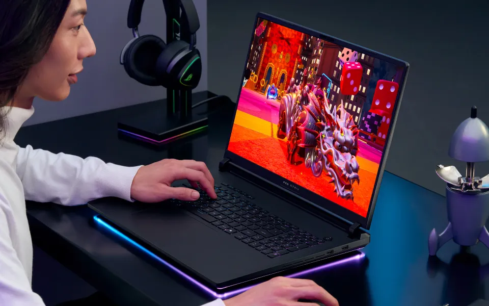 A person sitting at a desk with an ROG Strix laptop, playing a brightly lit game with a dragon on screen. 