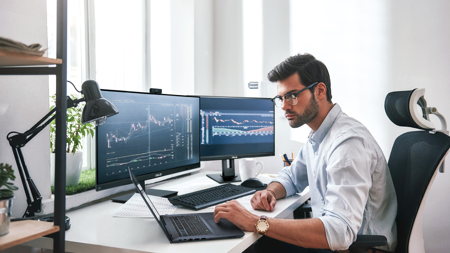 一名男子正在使用 ASUS ExpertBook 笔记本电脑监控股市实时信息，桌上有两台屏幕。