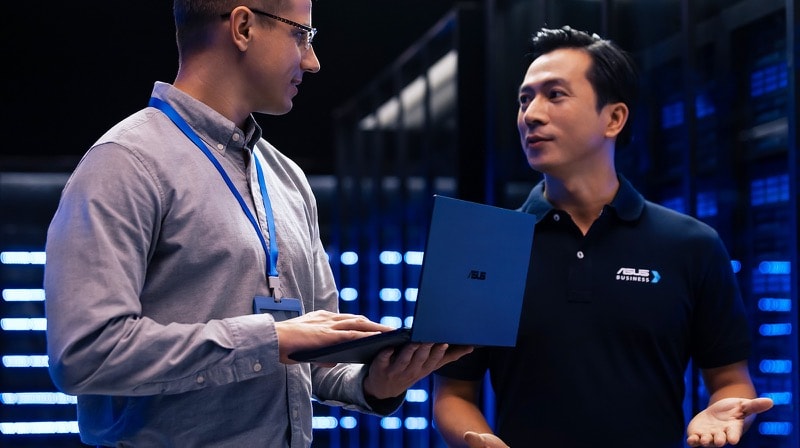 一位 IT 经理拿着一台 ASUS ExpertBook 笔电与一名 ASUS 业务人员在机房中交谈，业务人员穿着左胸有 ASUS Business 标志的 polo 衫。