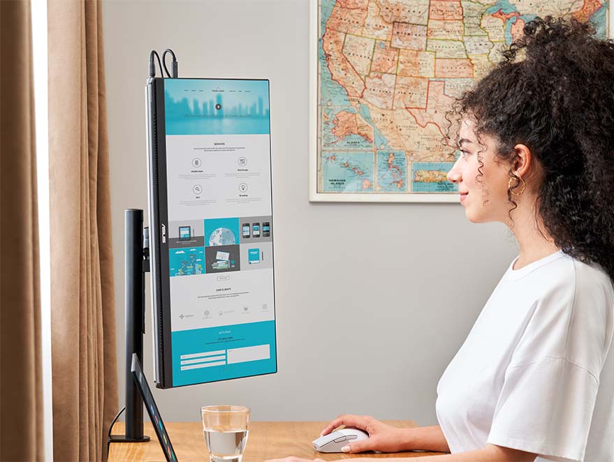 A woman stands working using a adjustment table, with a laptop and ZenScreen MB249C portable monitor set up, display long-from web page in portrait mode