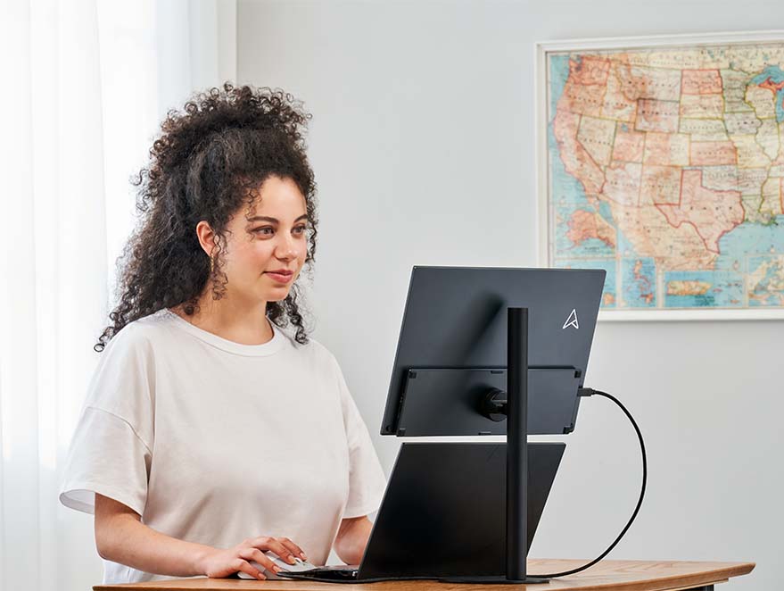 A woman stands working using an adjustment table, with a laptop and ZenScreen MB16QHG portable monitor mount on the MTS02SD stand, for comfortable working position