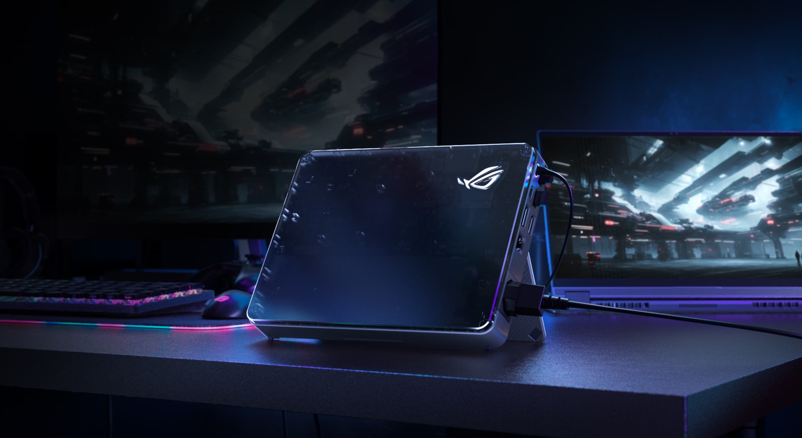 2025 XG Mobile 置于桌面中央的视图,前盖饰有 ROG 标志。背景中还有一面屏幕与 Flow X13。