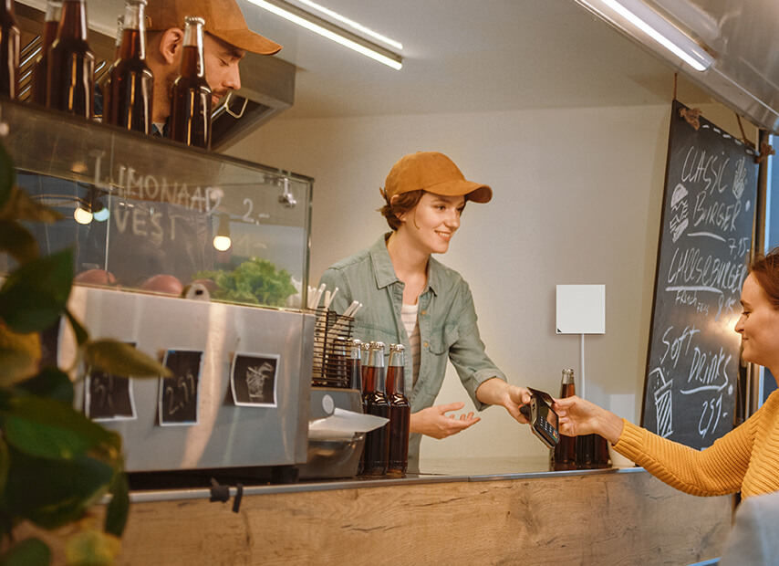 A food truck owner hands a card reader to a young female customer. The RT-AX57 Go router is wall-mounted inside the food truck.