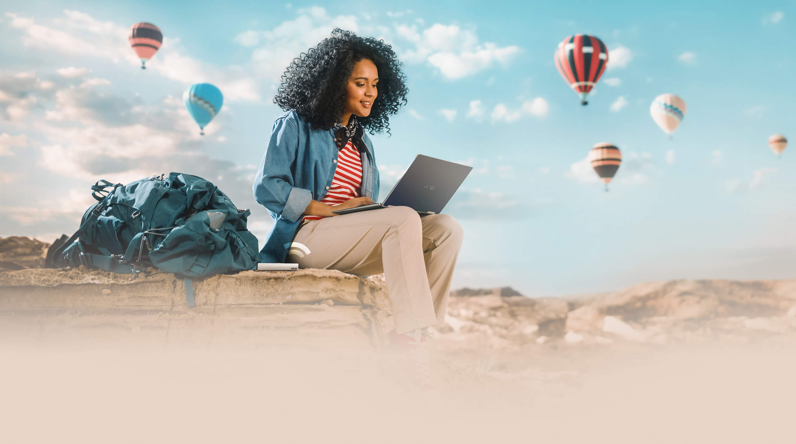 A young traveler seated outdoors on a rock, using a laptop with the RT-AX57 Go by side, against a backdrop of numerous hot air balloons dotting the blue sky.