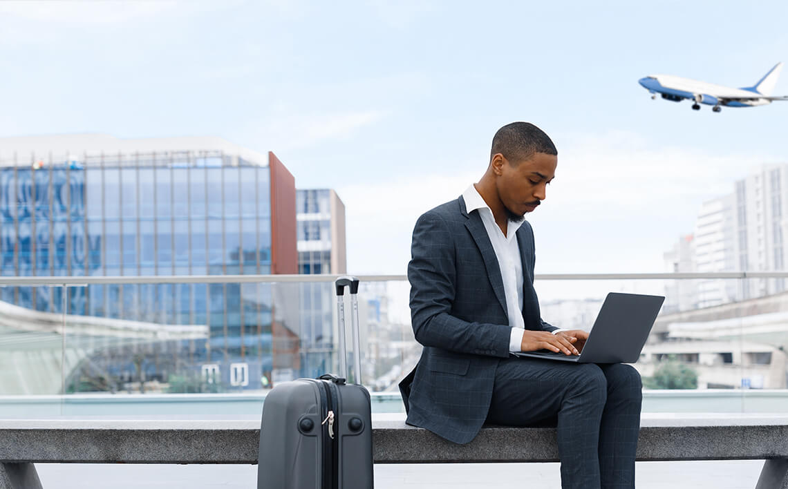 A businessman sits on a bench in the terminal hall, working on his laptop with a suitcase by his side, as a flight passes by in the sky overhead.