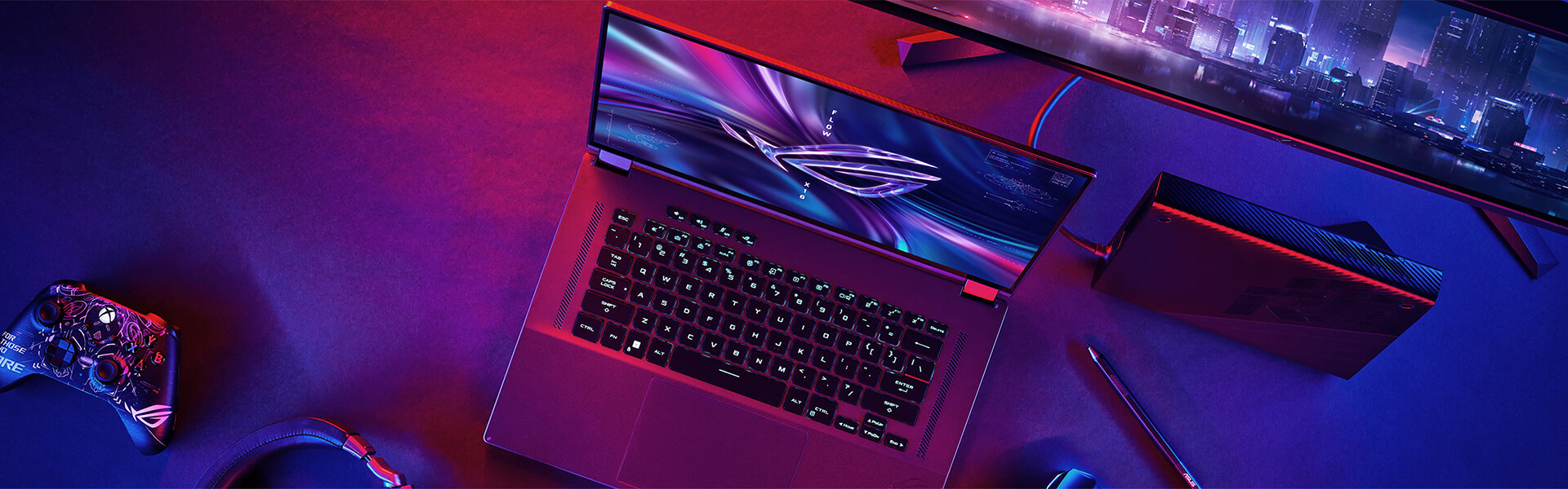 The ROG Flow X16 laptop sitting open on a desk next to a game controller, monitor, and XG Mobile external GPU