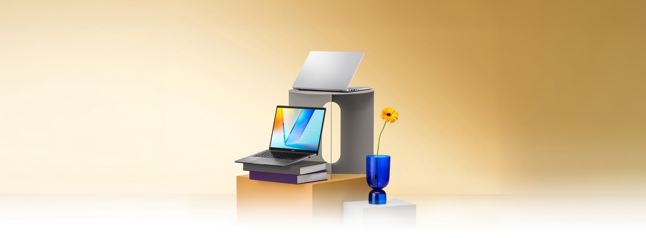 Two open laptops, one gray and one silver, are displayed on stacked gray and orange pedestals, with one resting on a pair of books. A single yellow flower in a blue glass vase stands nearby.