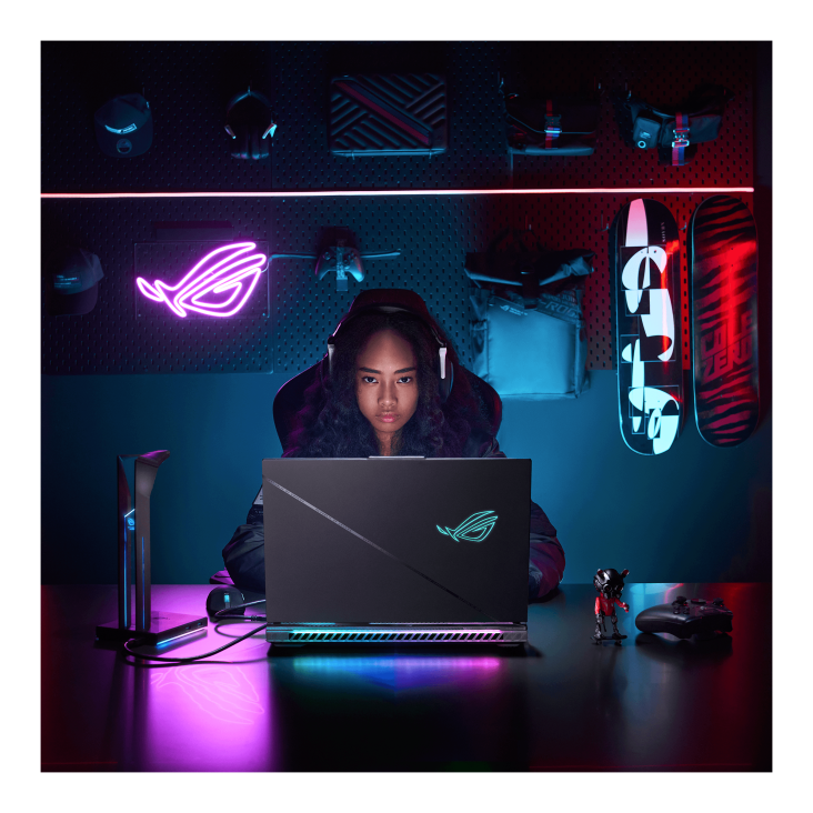 A dark room with purple lighting, a backpack, skateboards, and an ROG neon sign on the wall, with a gamer facing the camera and wearing a headset while using the ROG Strix SCAR laptop